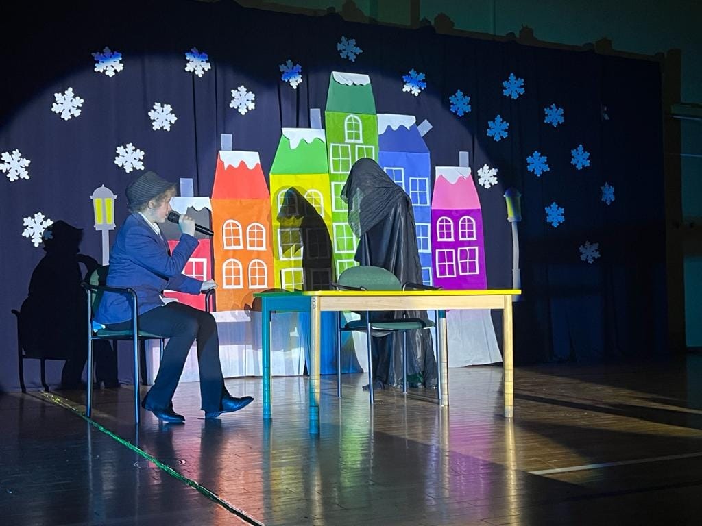 Na ilustracji przedstawienie ,,Opowieści Wigilijnej" w wykonaniu dzieci. Dwójka aktorów siedzi na krzesłach.