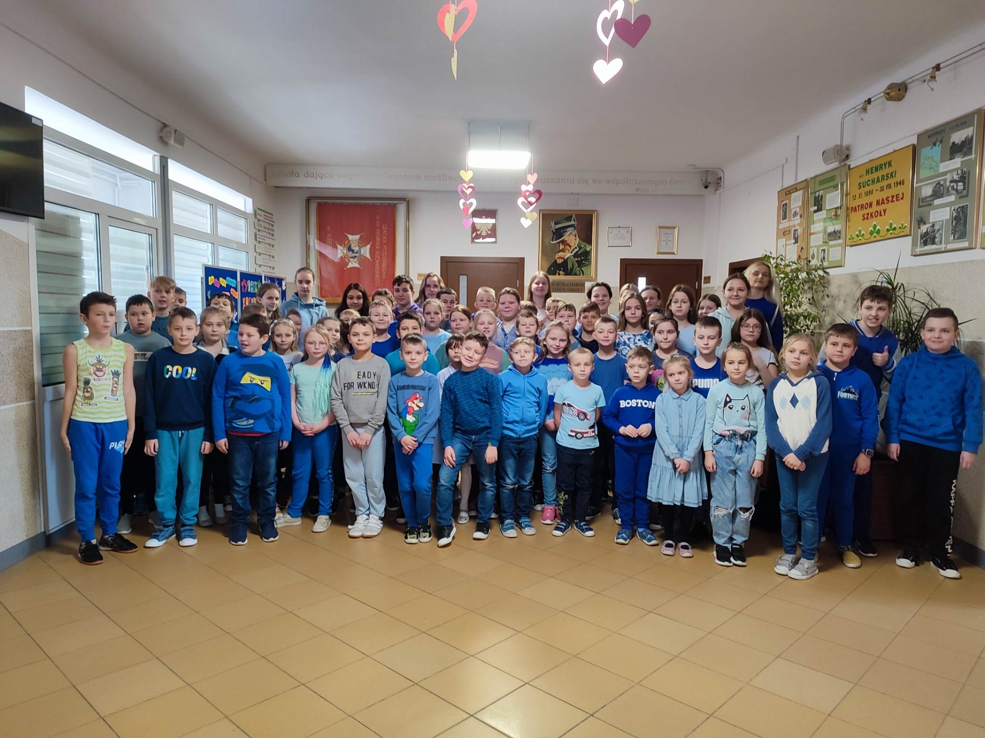 Uczniowie i nauczyciele na korytarzu szkolnym, ubrani w odcieniach niebieskiego.