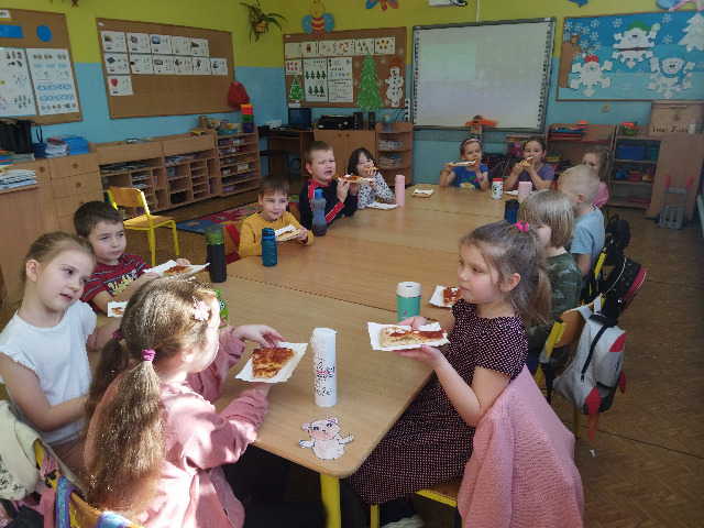 Na zdjęciu znajdują się dzieci z oddziału 6- latków.  Dzieci siedzą przy stoliku i jedzą pizzę. Zdjęcie zostało zrobione w sali przedszkolnej.