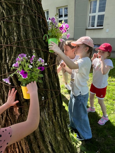 Widzimy kawałek pnia drzewa, na którym dzieci wieszają doniczki z zasadzonymi kwiatami.
