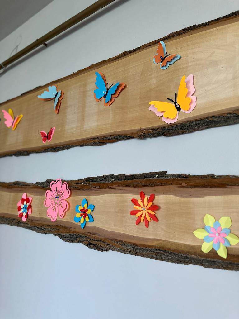 Zdjęcie drugie przedstawia dekorację wiosenną na świetlicy szkolnej. Na drewnianych deskach znajduje się wiosenna dekoracja, na dolnej wiszą kwiaty, a na górnej kolorowe motyle.