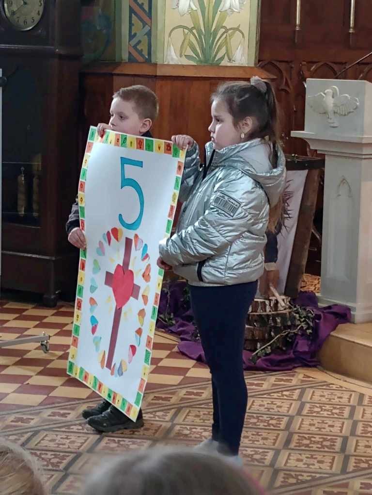 Na zdjęciu dziewczynka i chłopiec trzymają plakat przedstawiający piątą stację drogi krzyżowej.