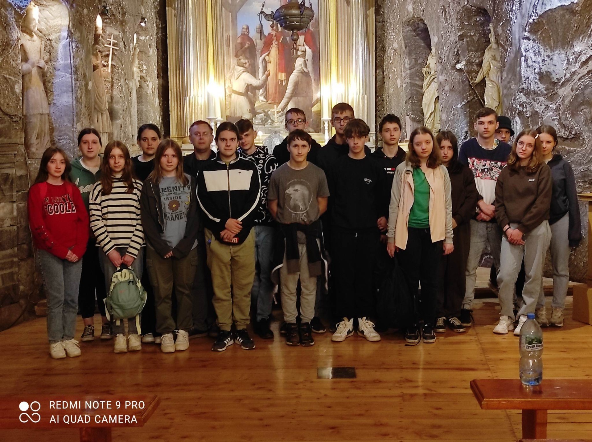 Zdjęcie przedstawia grupę dziewiętnastu uczniów i uczennic klas ósmych oraz nauczyciela. Uczniowie znajdują się w grocie kopalni w Bochni. Za nimi widoczny jest obraz oraz figury świętych, nad nimi lampka.