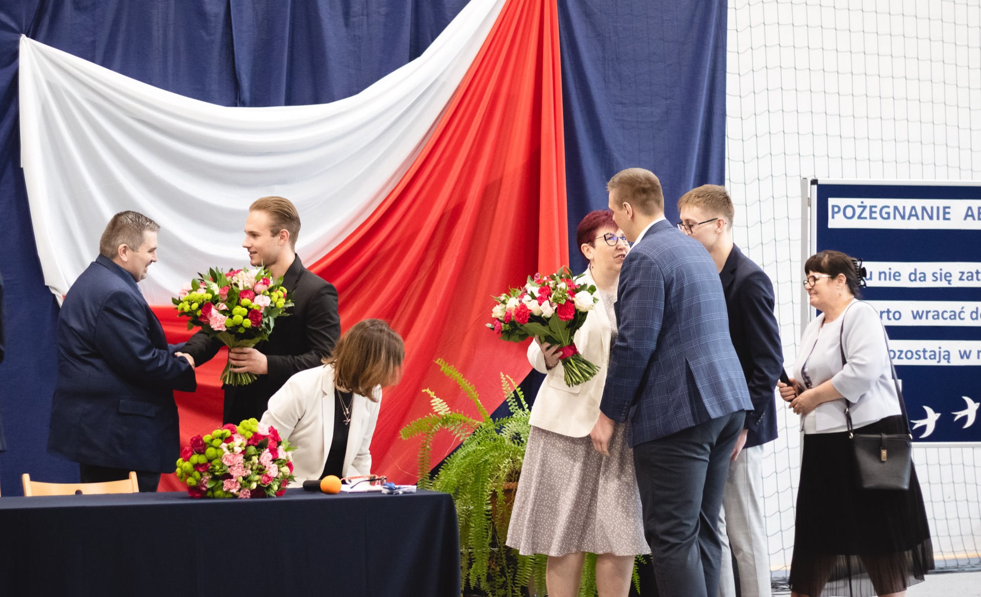 Absolwenci wręczają bukiety kwiatów dyrekcji oraz wychowawcom. W tle dekoracja biało-czerwona flaga na granatowym tle.