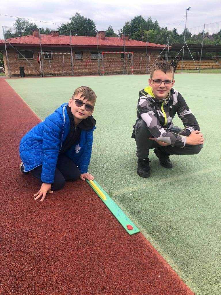 Zdjęcie przedstawia dwóch uczniów klasy czwartej na boisku szkolnym, którzy mierzą linijką długość boiska sportowego. 