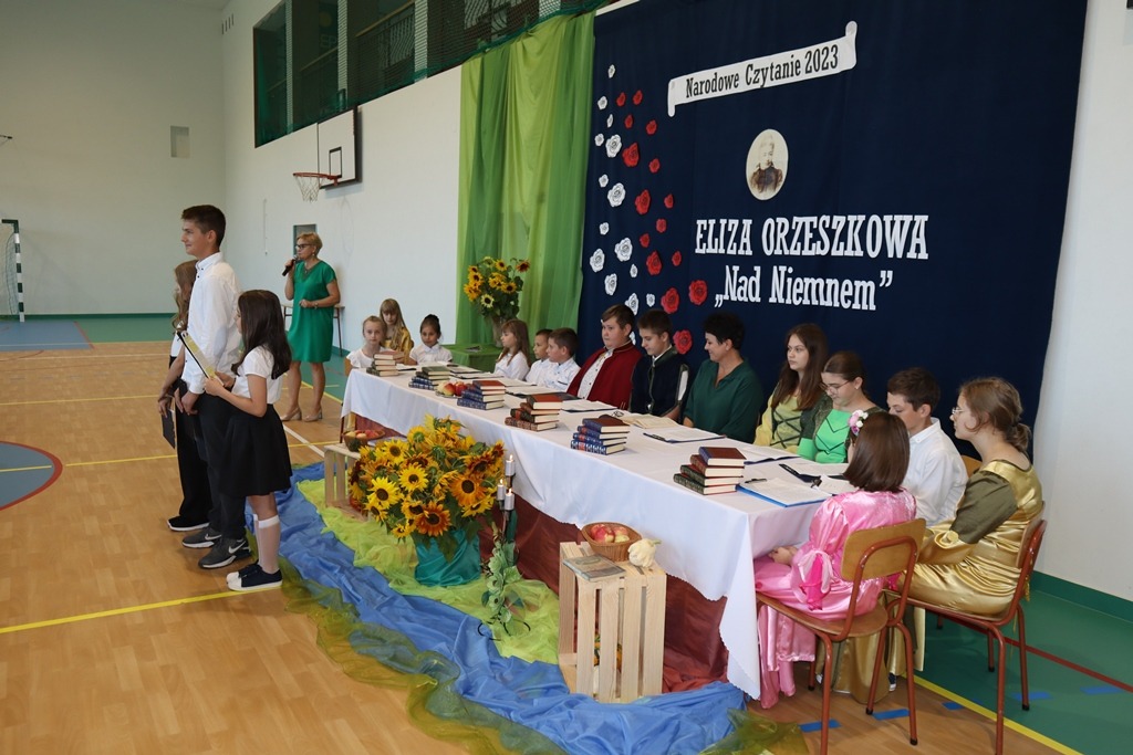 Fotografia przedstawia uczestników Narodowego czytania siedzących przy stole. Przed stołem stoi trójka uczniów zapowiadających czytających.
