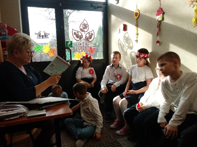 Przy ścianie po prawej stronie siedzą dzieci, pośrodku troje chłopców siedzi na dywanie. Po lewej stronie siedzi nauczycielka i trzyma książkę.