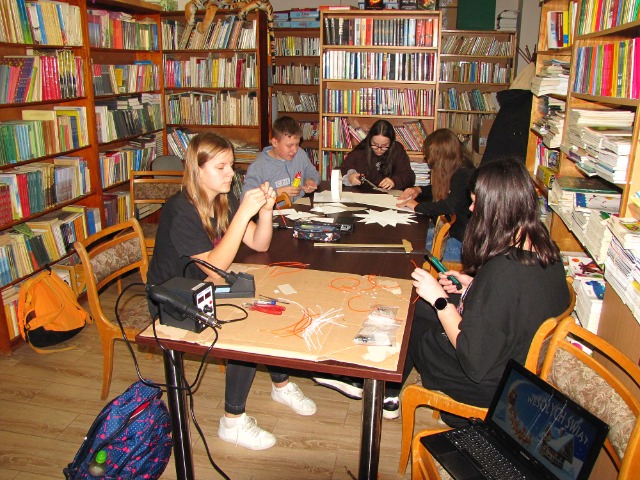 5 uczniów wykonuje model gwiazdy z kartonu modelarskiego i wykonują pierwsze połączenia elektryczne. Siedzą przy stole, w rękach trzymają części modelu, na stoliku stoi lutownica. Za ich plecami znajdują się regały z książkami. 