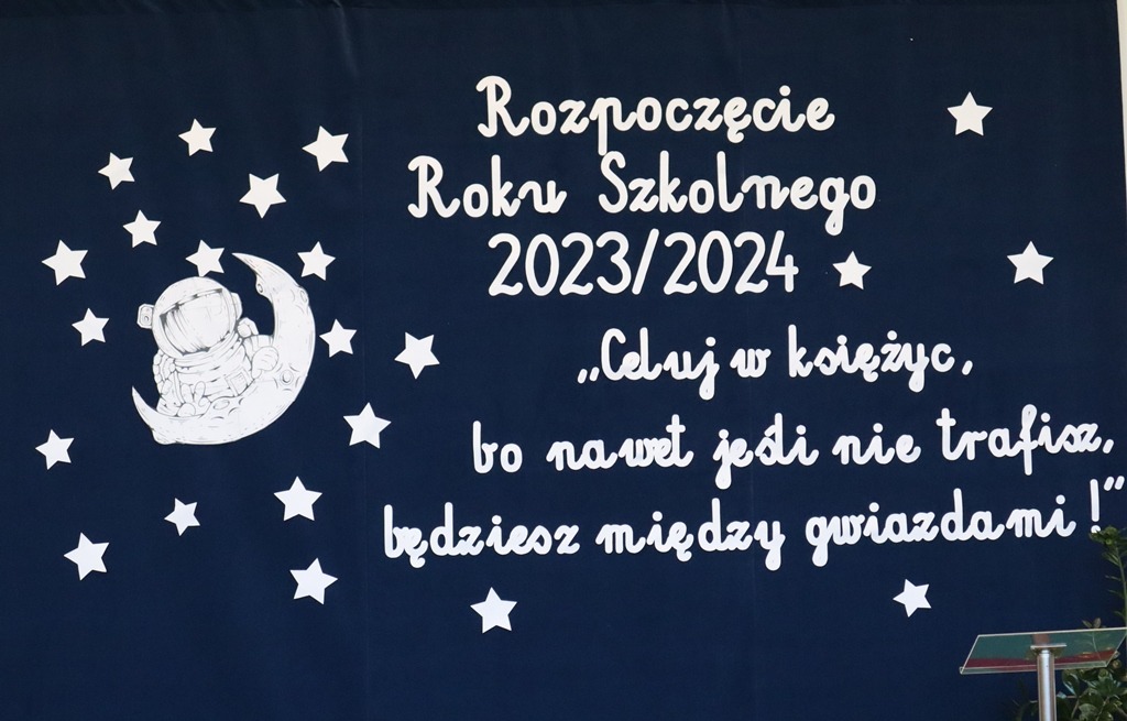 Na fotografii widzimy dekorację sali gimnastycznej wraz z hasłem Rozpoczęcie roku szkolnego 2023/2024 i cytat "Celuj w księżyc, bo nawet jeśli nie trafisz będziesz między gwiazdami!". Wokół napisu znajdują się gwiazdy. Z lewej strony napisu jest kosmonauta na księżycu.