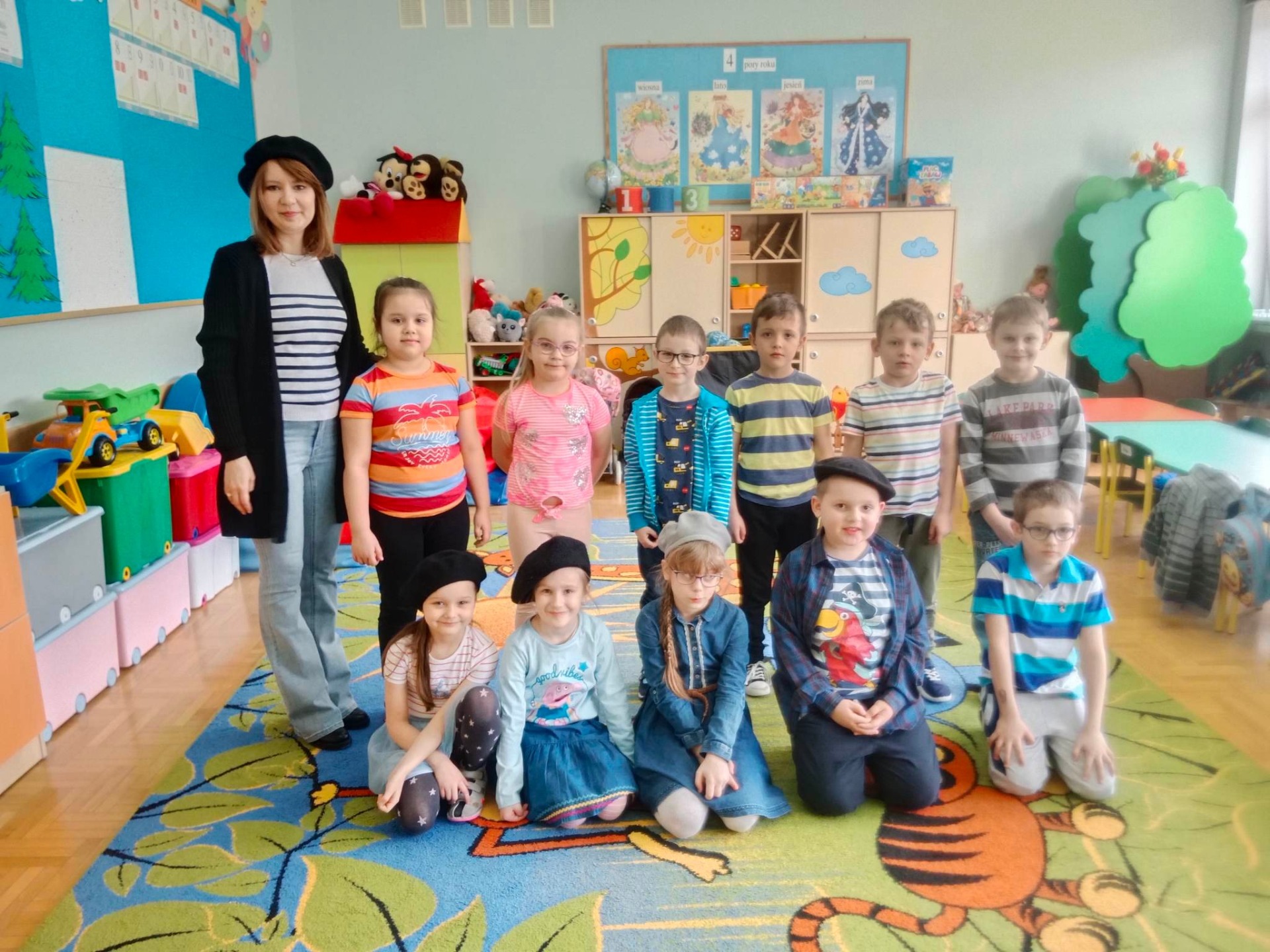 Piąte zdjęcie przedstawia nauczycielkę  i jedenastu uczniów klasy zero b, pięć dziewczynek i sześciu chłopców. Na pierwszym planie na kolorowym dywanie siedzi trzy dziewczynki i dwóch chłopców. Za nimi stoi nauczycielka, dwie dziewczynki i czterech chłopców. W tle kolorowe mebelki, zabawki i gazetki klasowe.