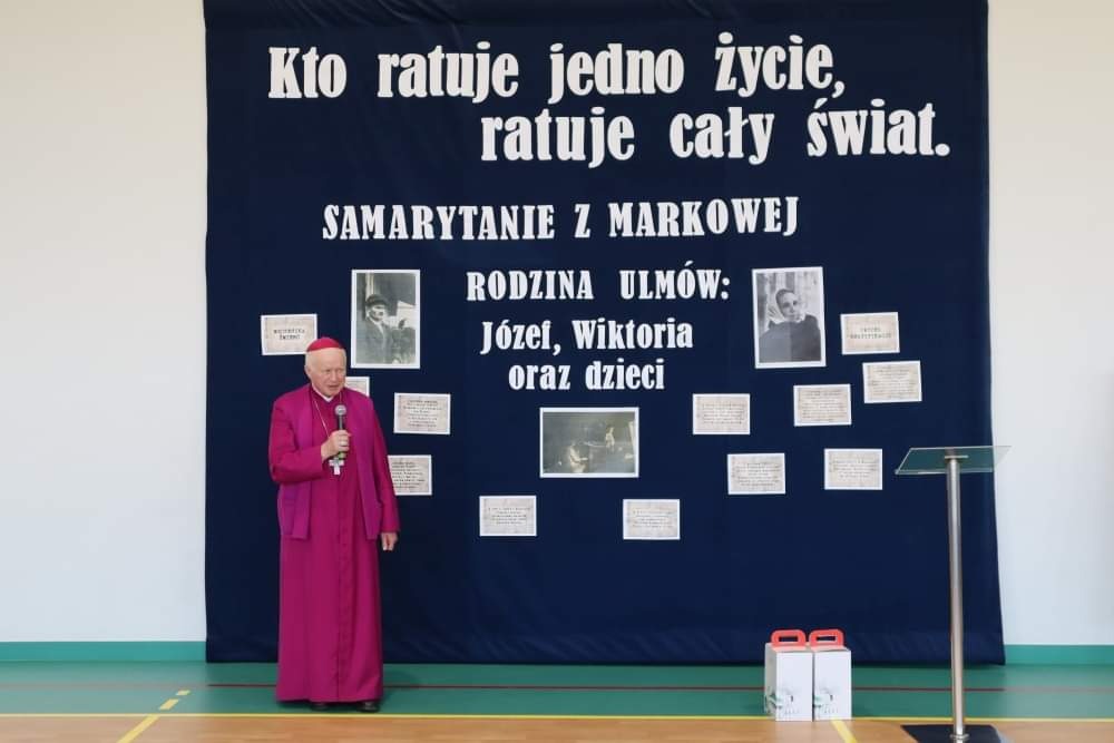 Na trzecim zdjęciu widzimy Arcybiskupa Adama Szala. W prawej ręce trzyma mikrofon. Za nim widać dekorację poświęconą rodzinie Ulmów - granatowe płótno z białymi napisami i zdjęciami. Po prawej stronie jest mównica, obok niej stoją dwa tekturowe białe pudełka z czerwonymi uchwytami.