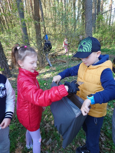 Uczniowie podczas sprzątania lasu. Na pierwszym planie uczeń trzyma worek na śmieci, a uczennica wrzuca do worka śmieci.

