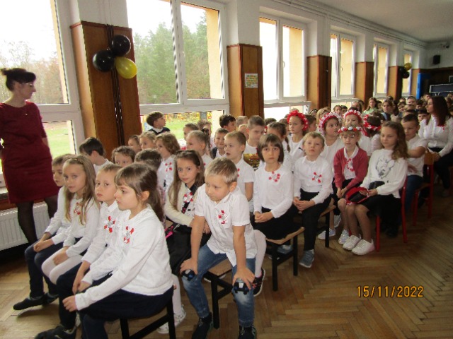 Korytarz szkolny. Uczniowie siedzą na ławeczkach ubrani w białe bluzki, do których mają przypięte biało -czerwone kotyliony.