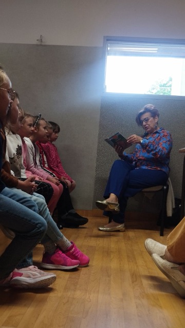 siedem dziewczynek siedzących bokiem wpatruje się w czytającą kobietę, która siedzi przed nimi na krześle, trzymając przed sobą książkę.