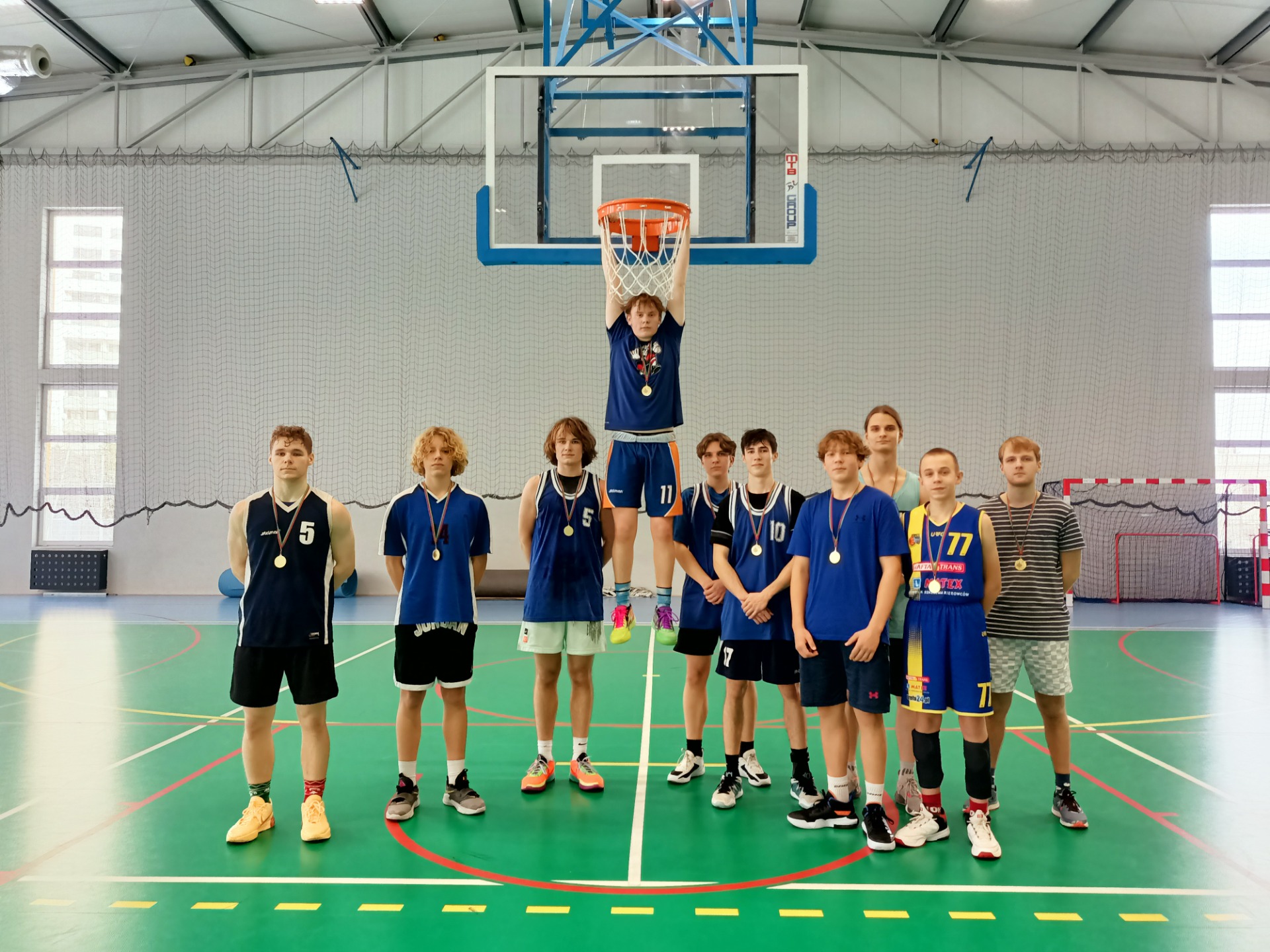 Zwycięska drużyna w niebieskich koszulkach z medalami  pozuje na sali gimnastycznej pod koszem do koszykówki. 