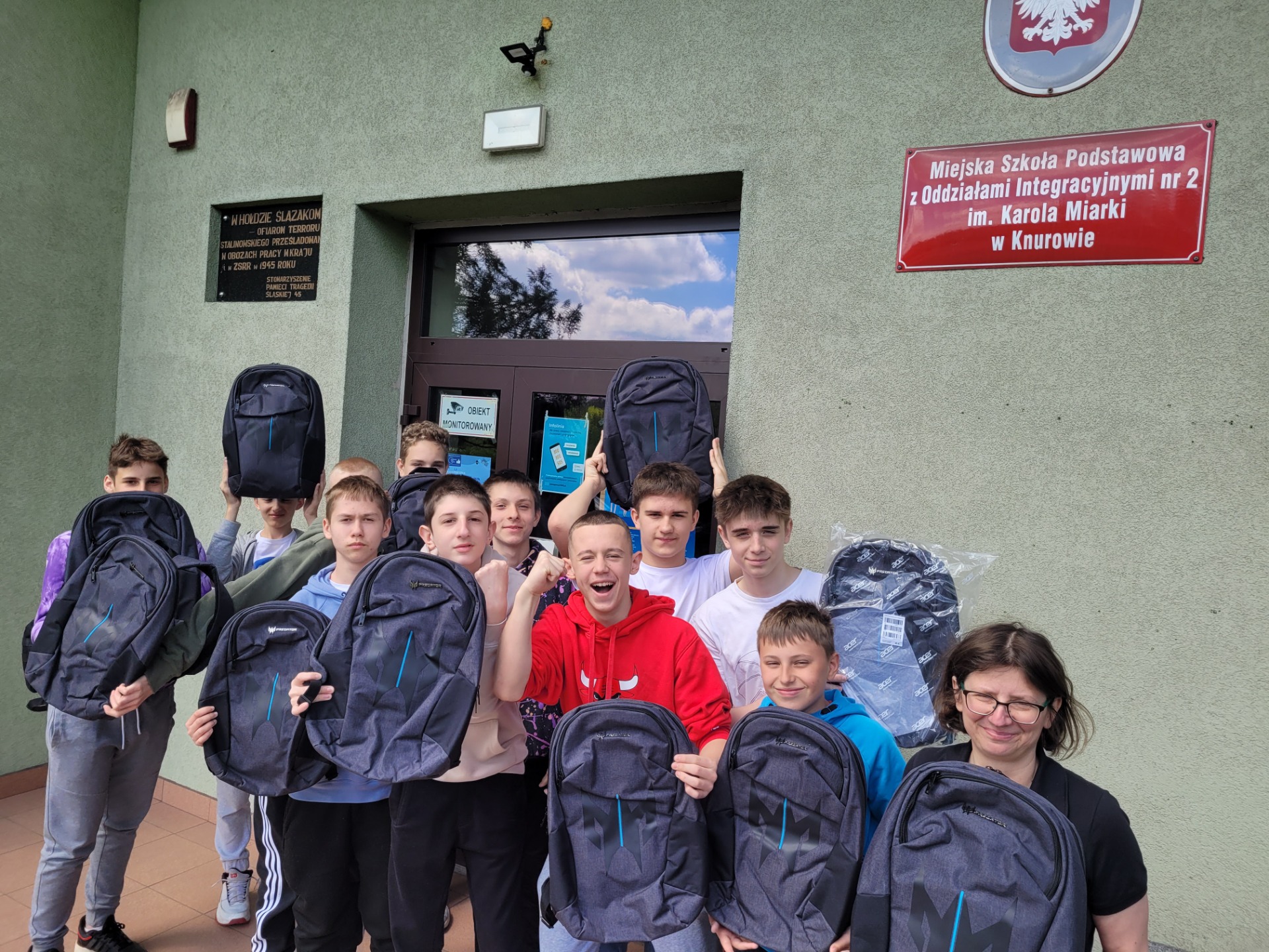 Grupa 10 uczniów wraz z nauczycielką trzymając plecaki uśmiechają się (krzyczą z radości) do zdjęcia przed budynkiem szkoły.