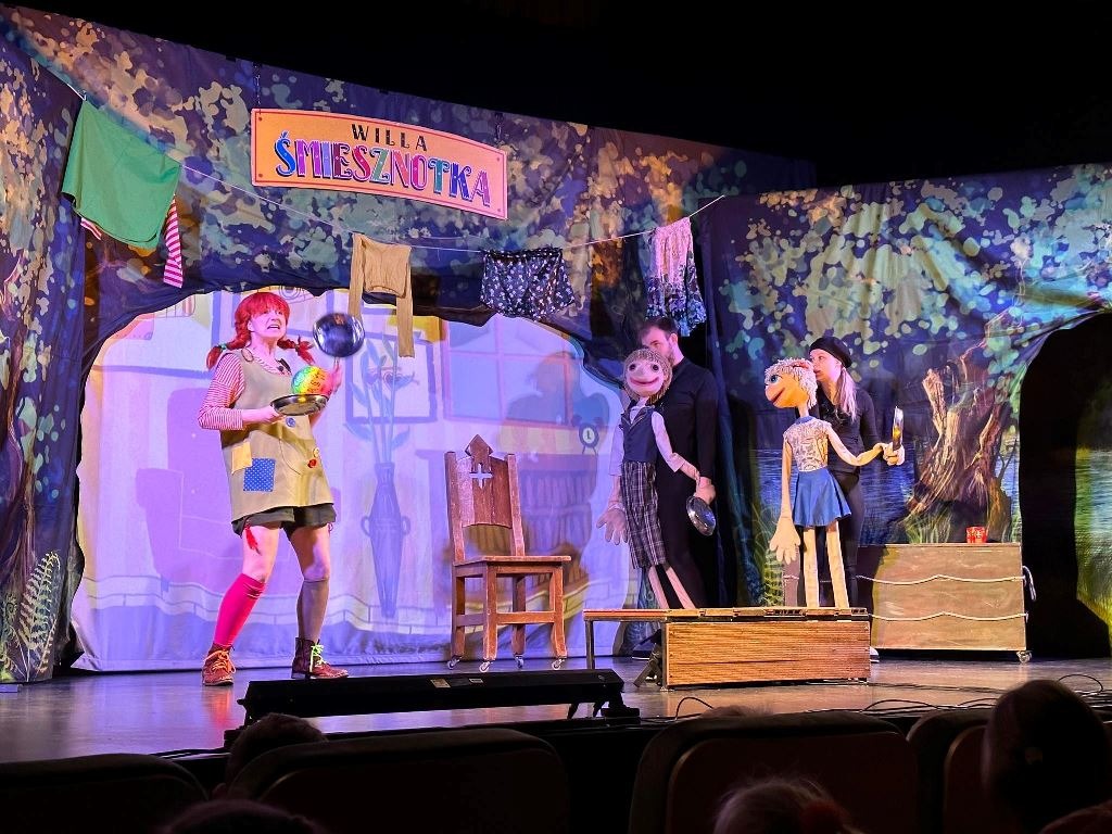 Na zdjęciu znajduje się trzech aktorów którzy stoją na scenie. Jedna z aktorek to dziewczyna o rudych włosach uczesana w dwa warkocze. W dłoniach trzyma patelnie. Dwaj aktorzy ubrani są na czarno i trzymają w dłoniach kukiełki. Na środki sceny znajduje się drewniane krzesło. Z tylu sceny umieszczona jest scenografia która przedstawia dom o nazwie Śmiesznotka