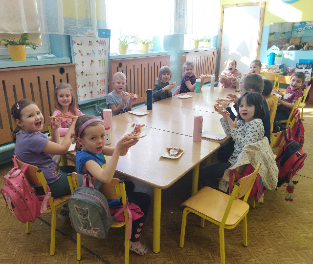 Na zdjęciu znajdują się dzieci z oddziału 6- latków.  Dzieci siedzą przy stoliku i jedzą pizzę. Zdjęcie zostało zrobione w sali przedszkolnej.