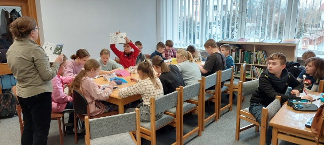 19 uczniów siedzi przy stole, rozwiązują zadania. Jedne uczeń trzyma w górze książkę. 